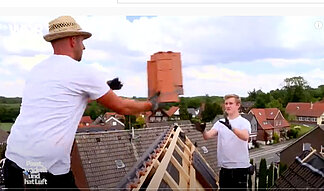 Zwei Dachdecker auf einem Dach werfen sich Dachpfannen zu.
